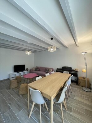Una sala de estar moderna con una mesa de comedor de madera, sillas blancas, un sofá, un televisor y un piano.