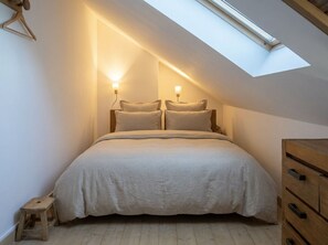 Ein Schlafzimmer mit schrägen Wänden, einem Dachfenster, einem Bett mit zwei Kissen, einem Holzschrank und einem kleinen Hocker.