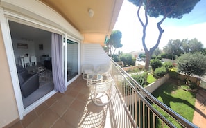 Un balcon avec une porte-fenêtre coulissante, un coin repas avec des chaises et une table, et une vue sur un jardin verdoyant et des voitures garées.