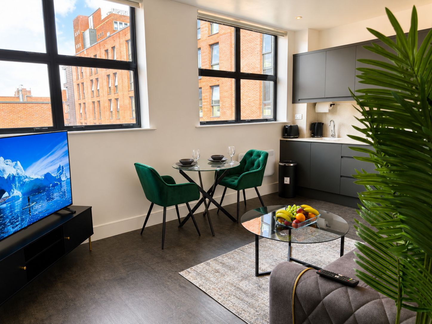 A modern living room with a flat-screen TV, green chairs, a glass coffee table with fruit, and a kitchen area with a TV and appliances.