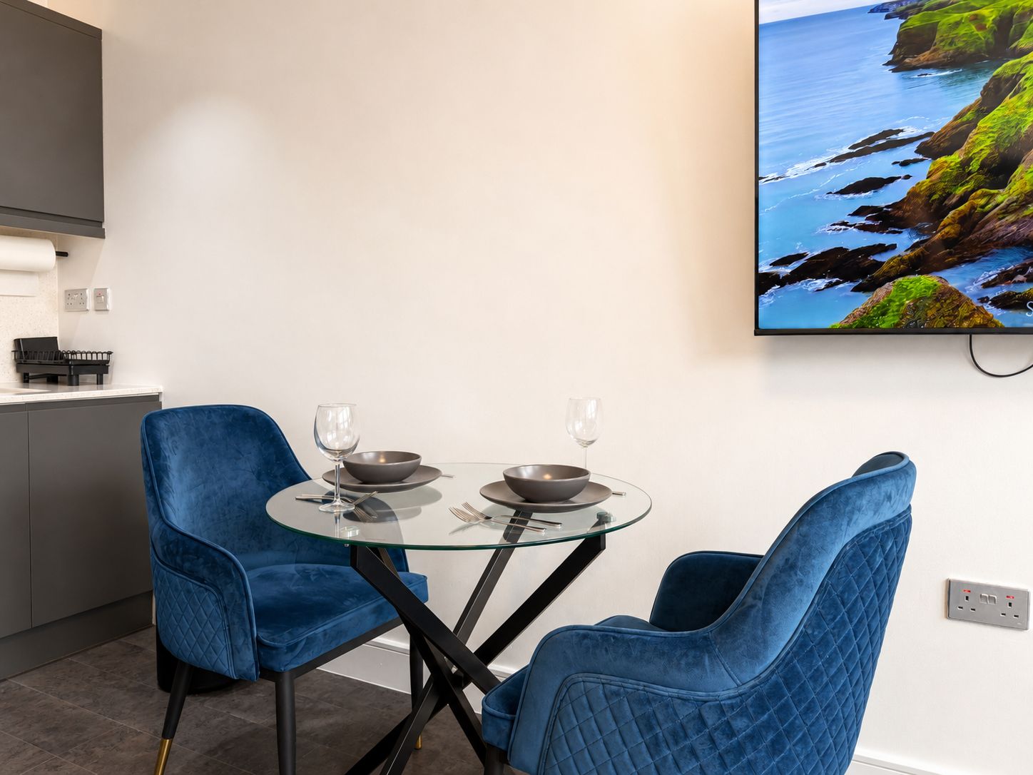 Uma área de refeições com duas cadeiras azuis, uma mesa de vidro e uma pintura de uma paisagem costeira.
