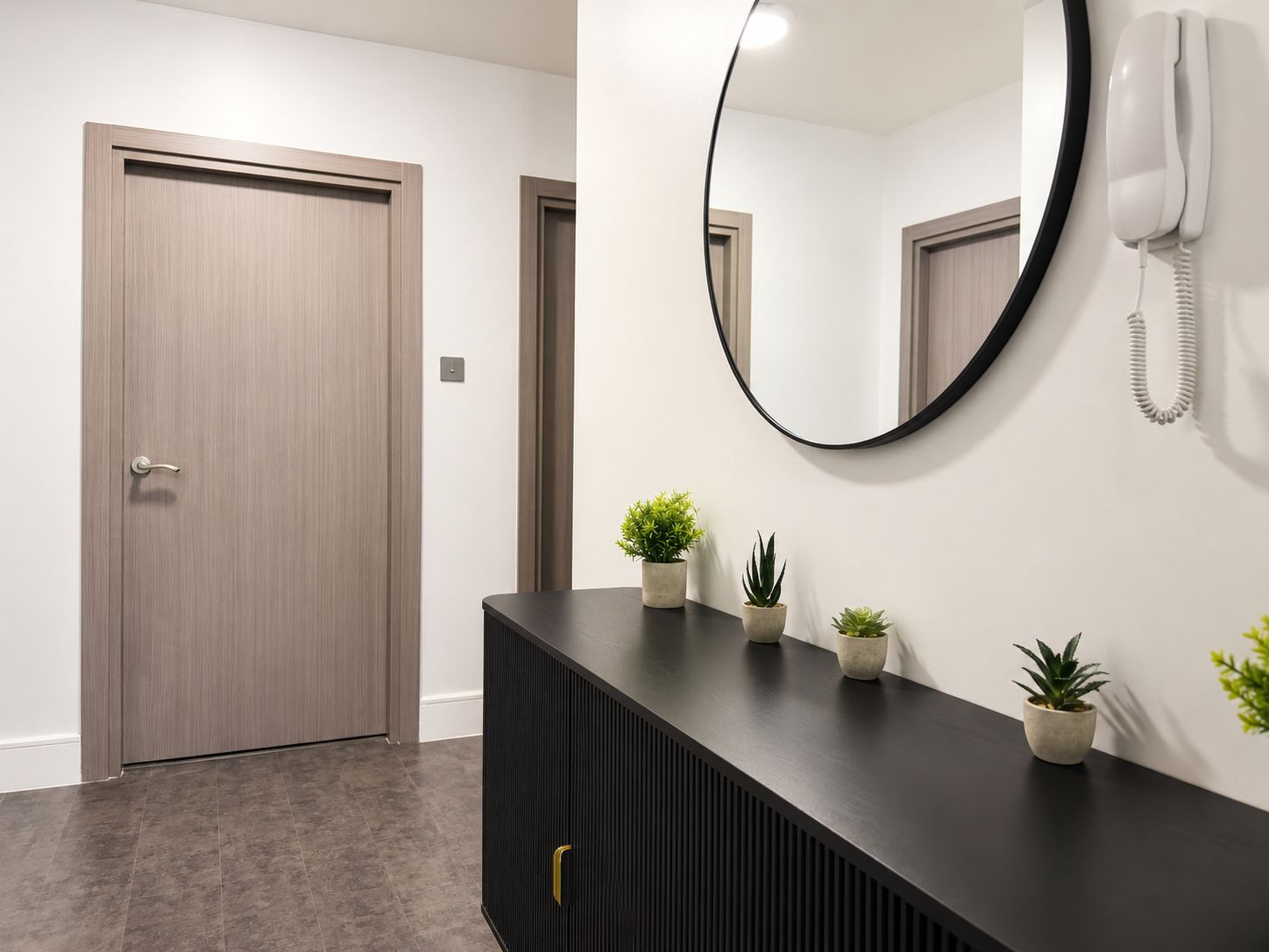 A hallway with a wooden door, a black console with plants, and a round mirror with a wall-mounted phone.