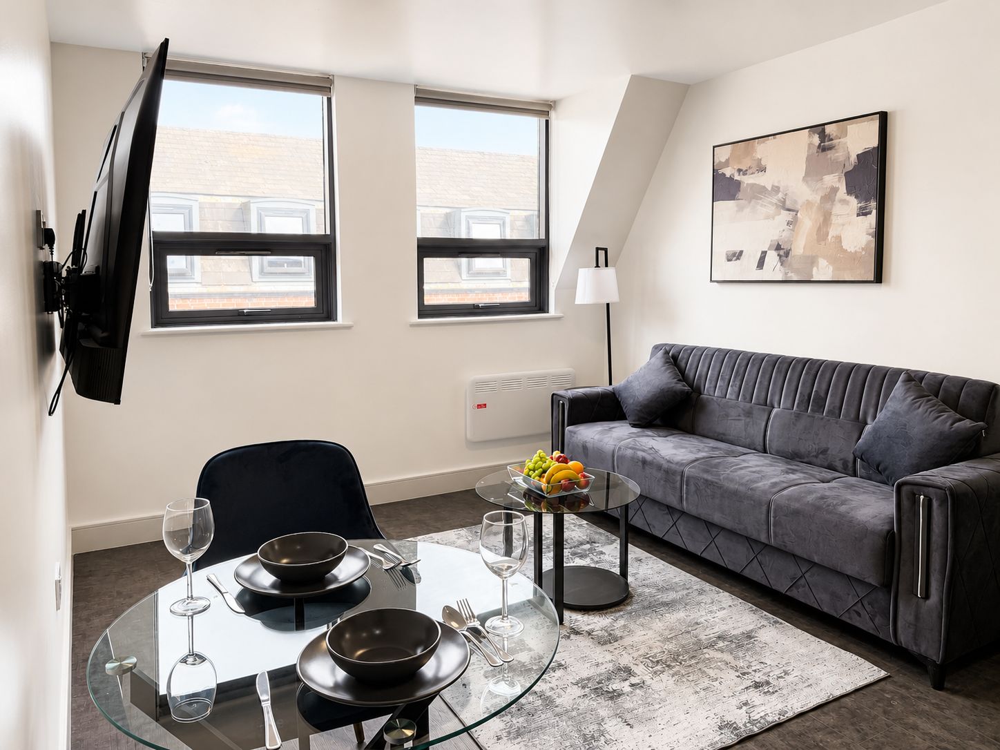 A modern living room with a glass dining table, a grey sofa, and a large window.