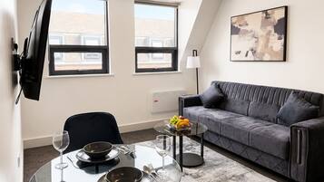 A modern living room with a glass dining table, a grey sofa, and a large window.