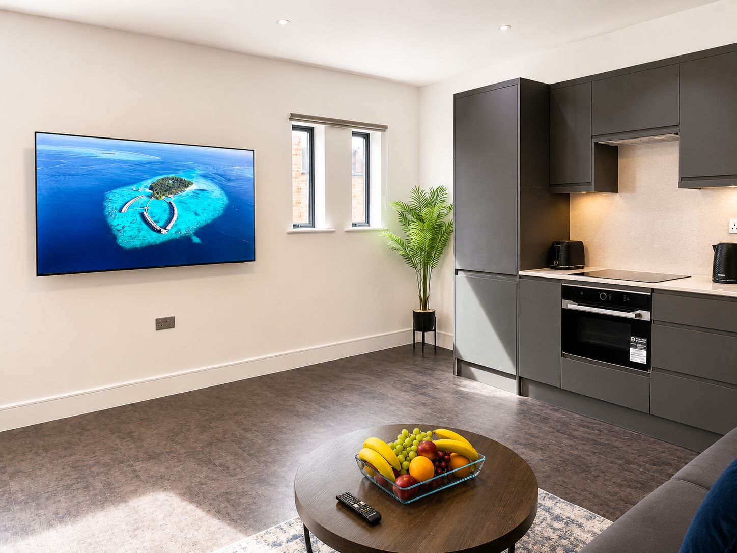 A modern living room with a flat-screen TV, a round coffee table with fruit, a sofa, and a kitchen area with dark cabinets and appliances.