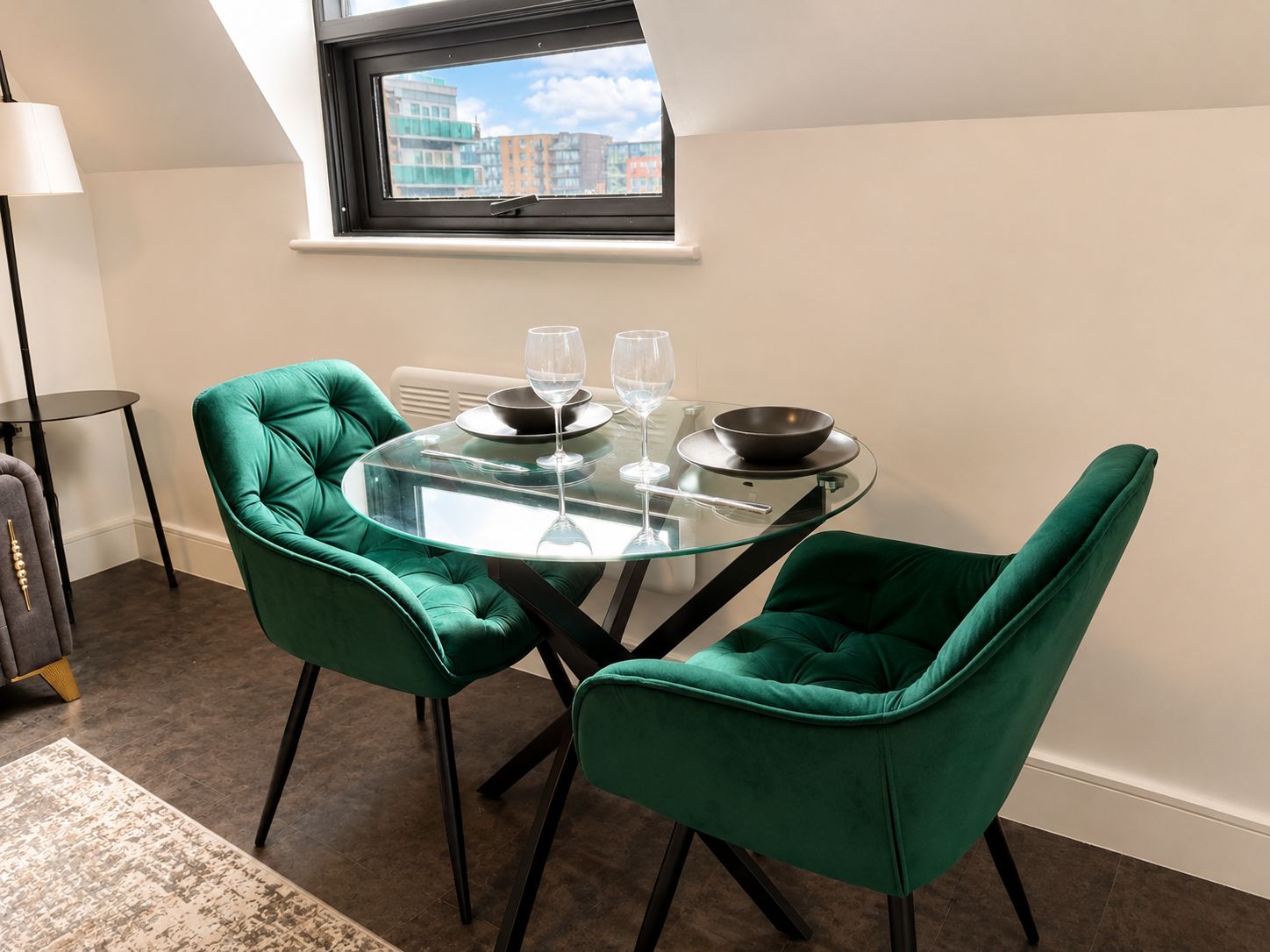A dining area with a glass table, two green upholstered chairs, and a window with a view of buildings.