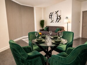 Ein Essbereich mit einem runden Glastisch, grünen Sitzmöbeln und einem braunen Sofa im Hintergrund.