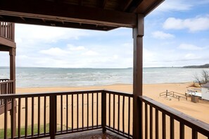 A beach view from a wooden deck with a railing, overlooking the ocean and a sandy beach. - Tidal Retreat 217 | Balcony | Direct Beach Access | Near Downtown & TART Trail (Traverse City)