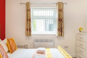 Een hotelkamer met een bed, een radiator, een raam met gordijnen, een witte dressoir en een stopcontact.