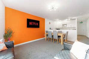 A modern living room with a large orange wall, a flat-screen TV, a dining area with a wooden table and chairs, and a kitchenette in the background. - Modern 5 Bed Flat in Thornaby Road For Big Groups (Thornaby)