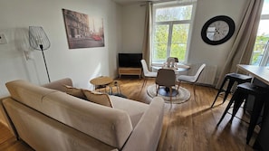Ein Wohnzimmer mit einer Couch, einem Fernseher, einer Essecke mit Stühlen und einem großen Fenster mit Vorhängen.