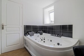 A modern bathroom with a large bathtub, a window above it, and a door to the left. - Le Nid Cosy 4, Balnéo 2 lits wifi (Tourcoing)