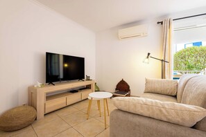 A modern living room with a flat-screen TV, a wooden entertainment unit, a round white table, a beige sofa, and a window with curtains. - Rated apartment with pool & quiet terrace (Valras-Plage)