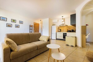 A living room with a sofa, two small tables, a kitchen with modern appliances, and framed pictures on the wall. - Rated apartment with pool & quiet terrace (Valras-Plage)