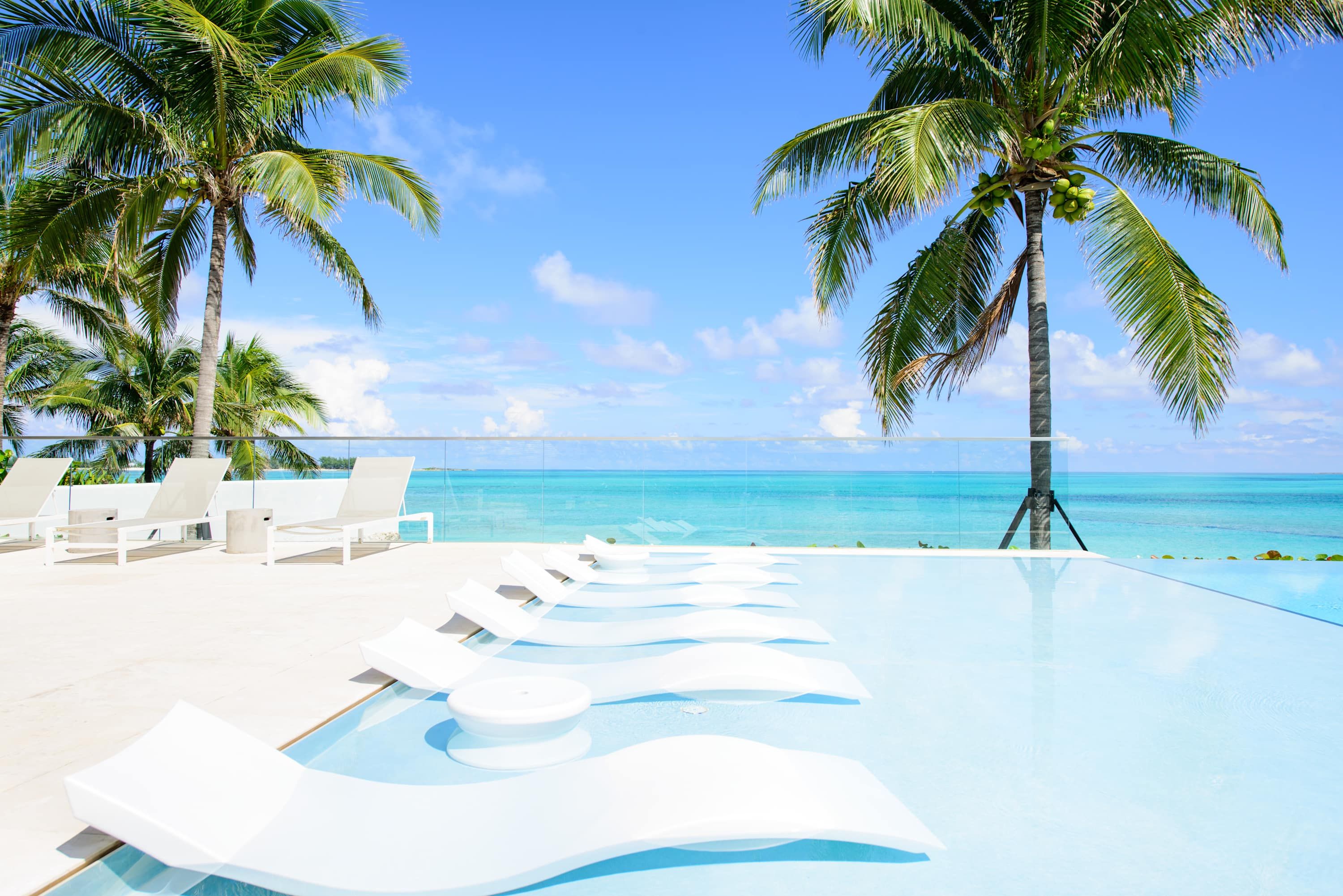 A poolside area with lounge chairs and palm trees, overlooking a clear blue sea.