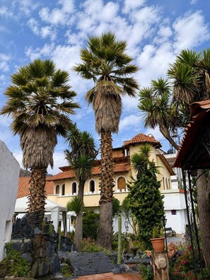 A courtyard with palm trees, a building with a tiled roof, and a stone wall.