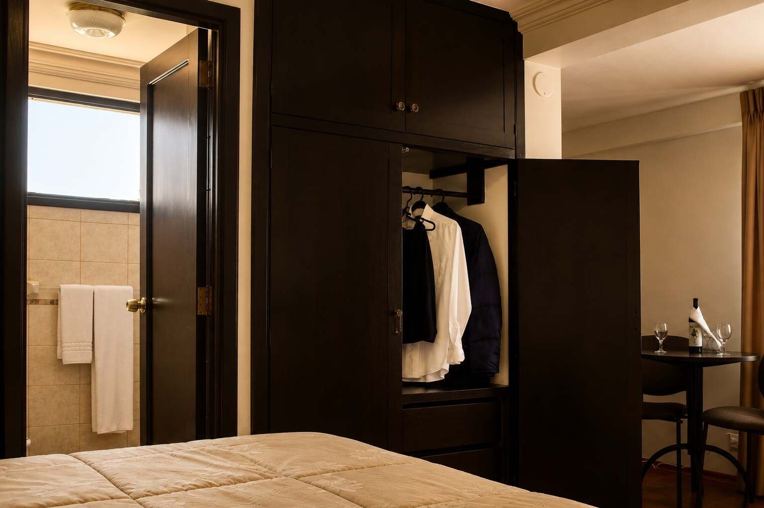 A hotel room with a bed, a wardrobe, a mirror, a small table with a bottle and glasses, and a towel rack.