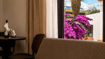 A room with a sofa, a table with a glass, and a window with a view of a palm tree and flowers.