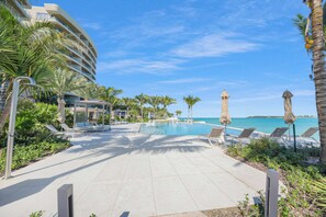 A resort pool area with lounge chairs and umbrellas, surrounded by palm trees and a clear view of the ocean. - Oceanfront Three-bedroom Condo with Infinity Pool (Nassau)