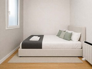 Un letto singolo con materasso bianco, coperta grigia e due cuscini, un tappeto grigio e un termosifone bianco.