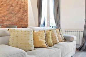 Un soggiorno con un divano bianco, cuscini beige e gialli, un muro in mattoni e una finestra con tende.
