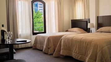 A hotel room with two beds, a round table, a chair, and a view of greenery through the window.