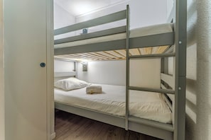 Ein Schlafraum mit Etagenbetten, Holzboden, weißen Wänden und einem kleinen Nachttisch mit Lampe.