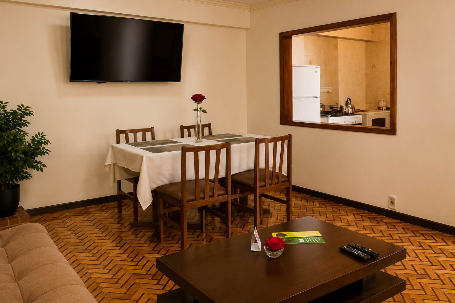 A room with a dining table set for four, a flat-screen TV mounted on the wall, a kitchen visible through a mirror, and a potted plant.