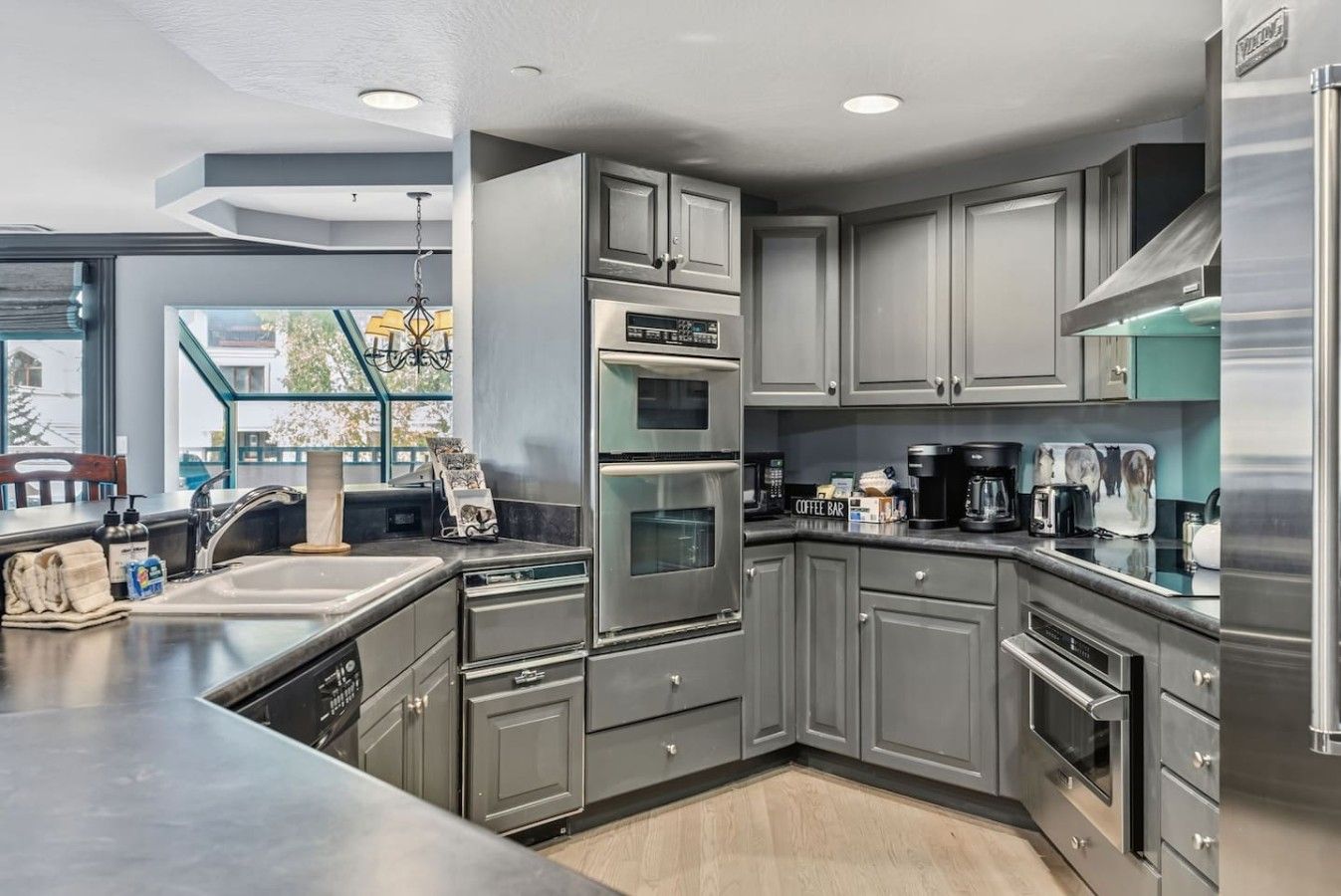 A modern kitchen with stainless steel appliances, grey cabinetry, and a large window providing natural light.