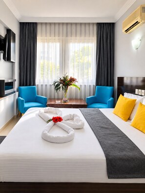 A hotel room with a bed, two blue armchairs, a wooden table with a vase of flowers, and a TV mounted on the wall. - Lara Palace Hotel (muratpaşa)