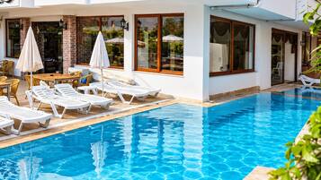 Una piscina con sillones de descanso y sombrillas, un muro de ladrillo y un edificio blanco con ventanas amplias.