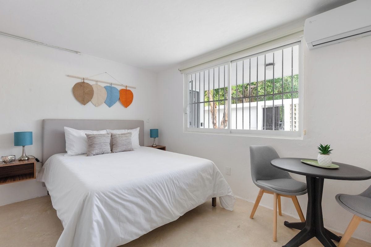Quarto com cama, mesas de cabeceira, janela com grades, uma cadeira e uma pequena mesa.