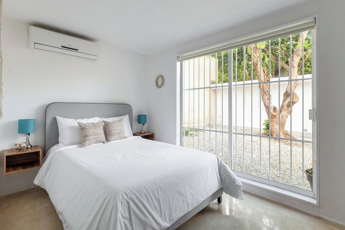 A bedroom with a bed, nightstands, a lamp, a window with a view of trees, and an air conditioning unit.