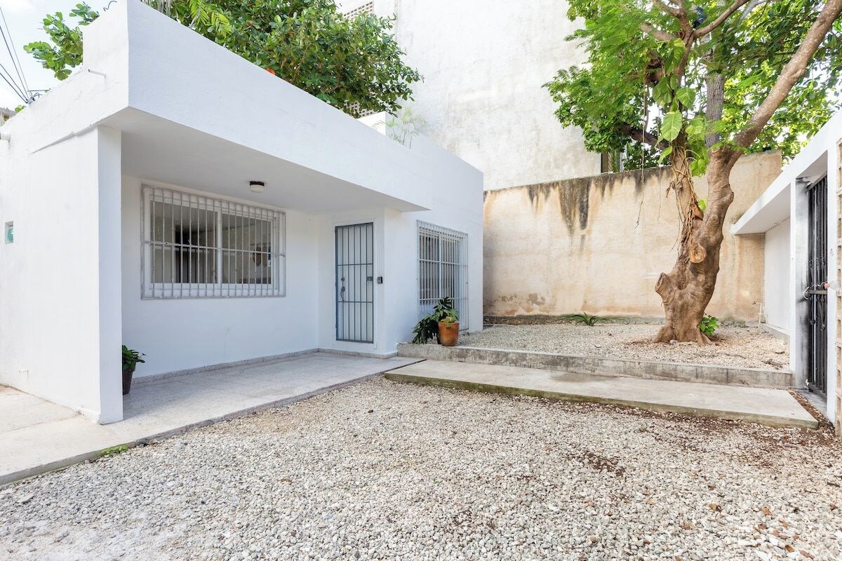 Um edifício branco com grades nas janelas, entrada com calçamento de brita e uma árvore no pátio.