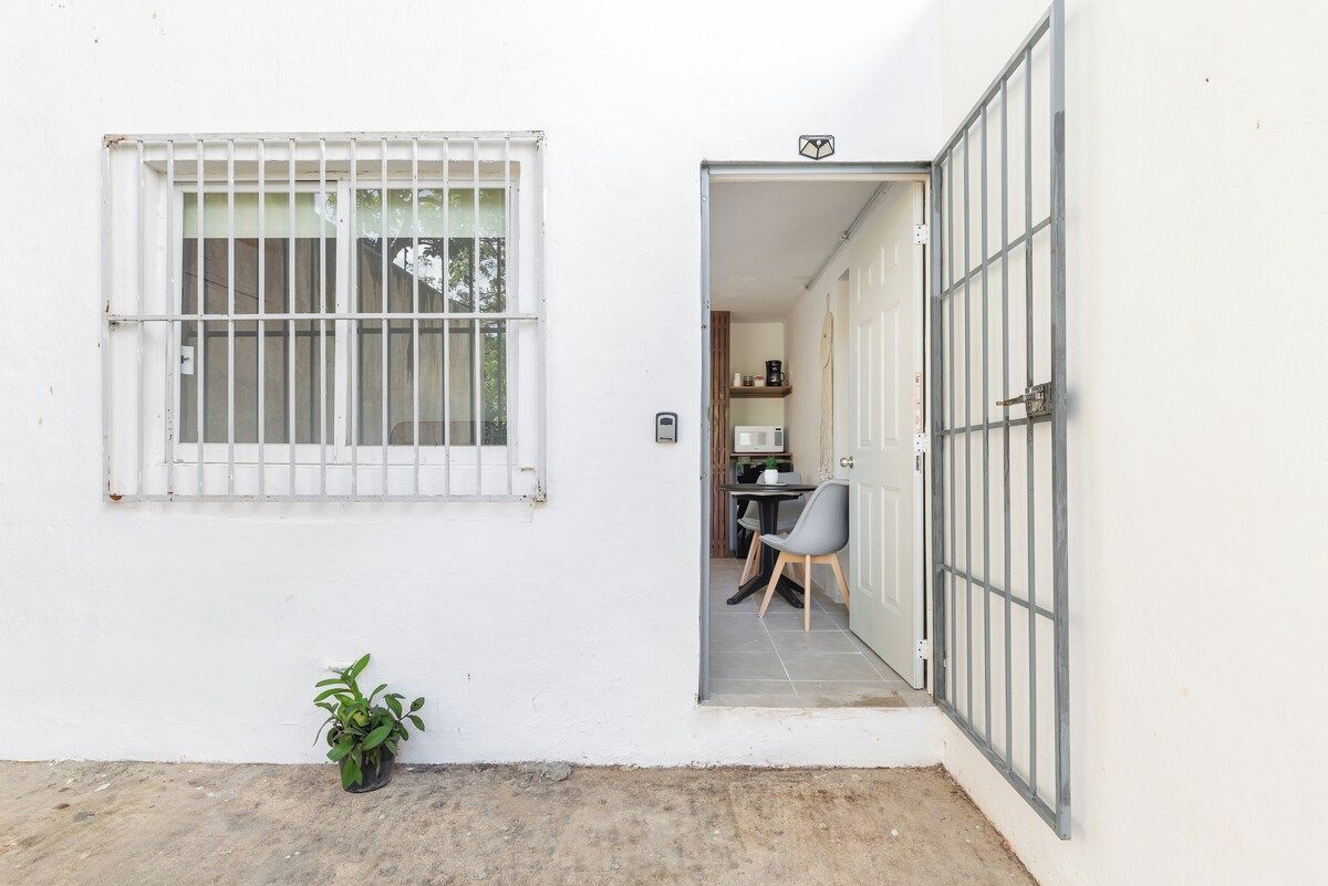 Paredes externas brancas com uma janela protegida por grades e uma porta que dá para um espaço interior com uma mesa e uma cadeira.