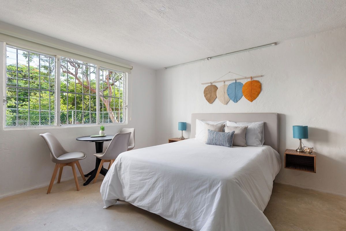 Un dormitorio con cama, dos sillas, una mesita y ventana con vista a árboles.