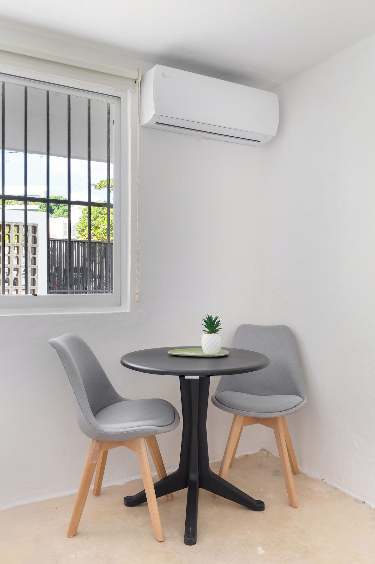 A small room with a round table and two chairs, a window with bars, and an air conditioning unit mounted on the wall.