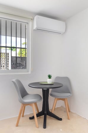 Quarto pequeno com mesa redonda e duas cadeiras, janela com grades e um ar condicionado fixado na parede.