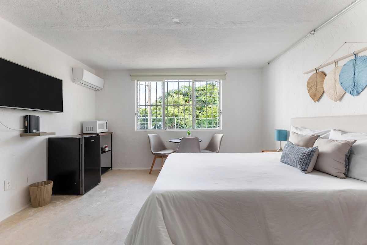 Quarto com cama, escrivaninha, cadeira, televisão e janela com vista para área verde.