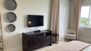 Quarto com televisão na parede, cómoda de madeira escura, mesa de apoio redonda, cadeira bege e janela grande com cortinas.
