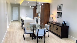 A modern kitchen with a central island, a dining area with a glass table, and a dark wooden cabinet. - 5th Floor Condo at ONE Cable Beach (Nassau)