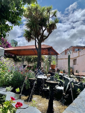 Ein Garten mit einem Brunnen, einer Palme und einem Gebäude mit rotem Dach im Hintergrund.