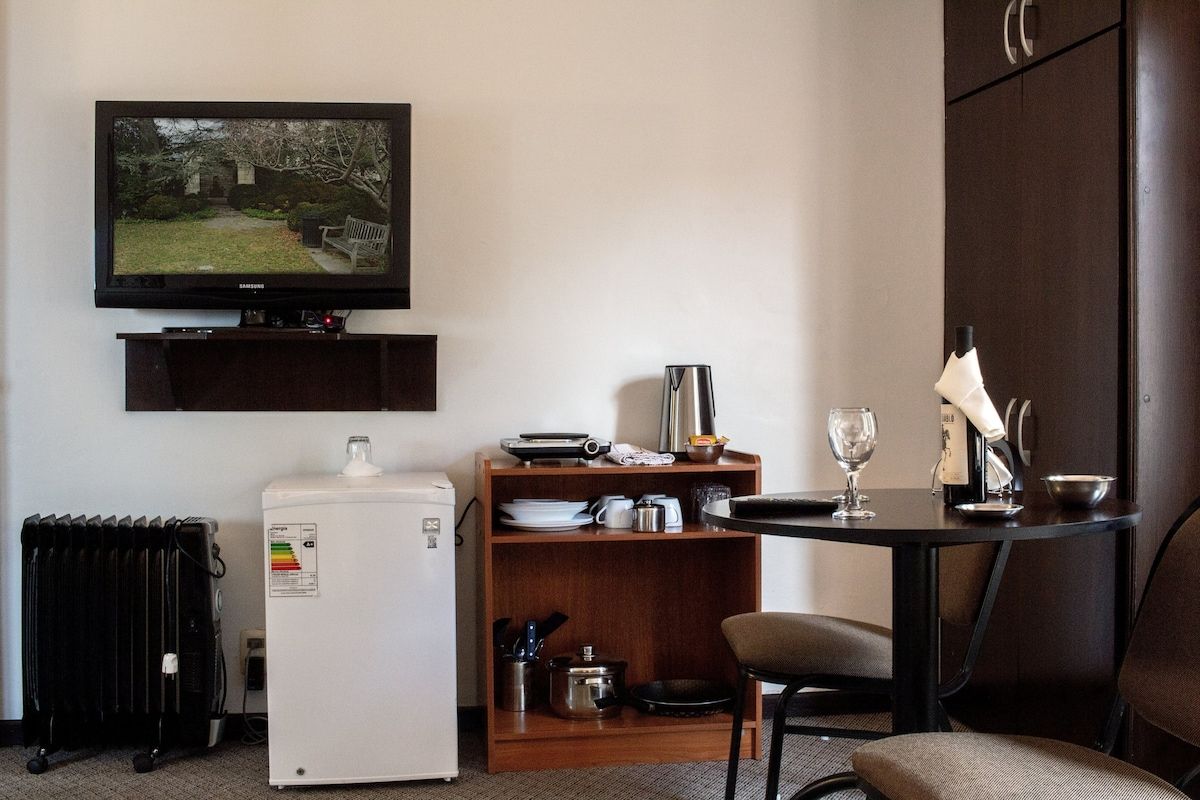 A compact hotel room with a television, a small refrigerator, a dining table with a chair, and a cabinet.