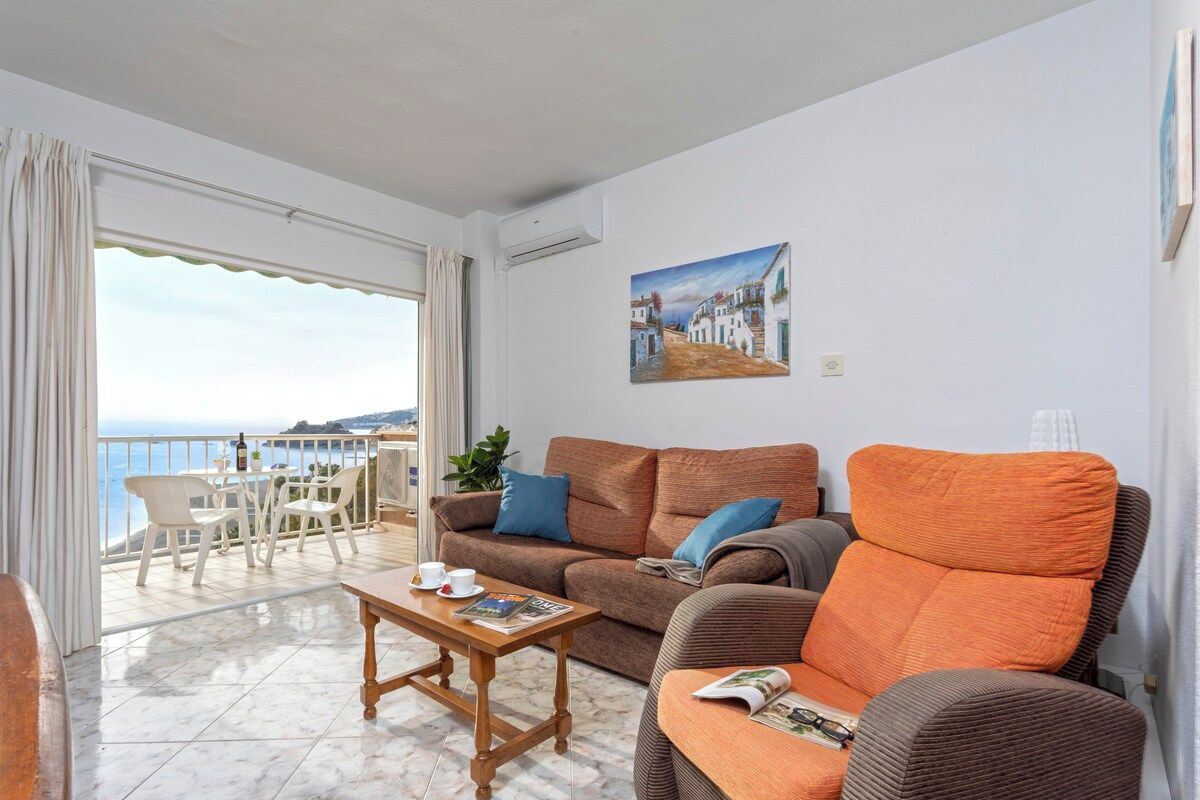 A living room with a sofa, armchair, and coffee table, a balcony with a view of the sea, and a painting on the wall.