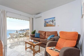 Un salon avec un canapé, un fauteuil et une table basse, un balcon avec vue sur la mer et un tableau accroché au mur.