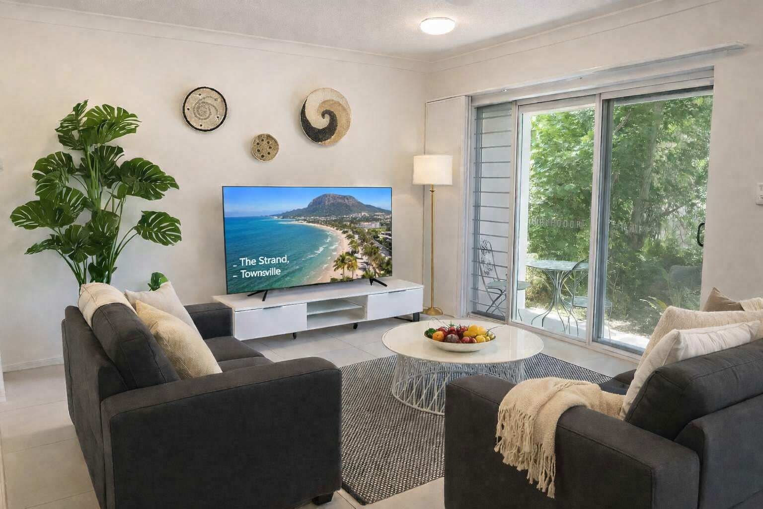 A modern living room with a large flat-screen TV, two grey sofas, a coffee table with fruit, and a large window with a view of a beach.