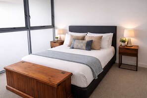 A neatly made bed with a grey throw blanket, two bedside tables with lamps, and a wooden bench. - Townsville CBD Stadium 2BR, 2BA w Pool, Spa & Parking (Townsville City)