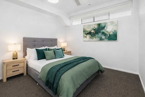 A bedroom with a bed, two bedside tables, a nightstand, a wall-mounted painting, and a ceiling fan. - CBD Balcony, Stadium & V8 Views & 2 Car Parks (Townsville City)