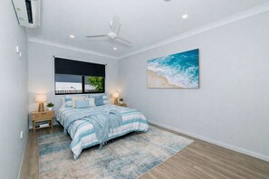 Un dormitorio con cama, ventilador de techo, ventana con vistas, un cuadro en la pared y una mesita de noche con lámpara.