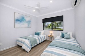 Quarto com duas camas, um ventilador de teto, janela com vista e um quadro na parede.